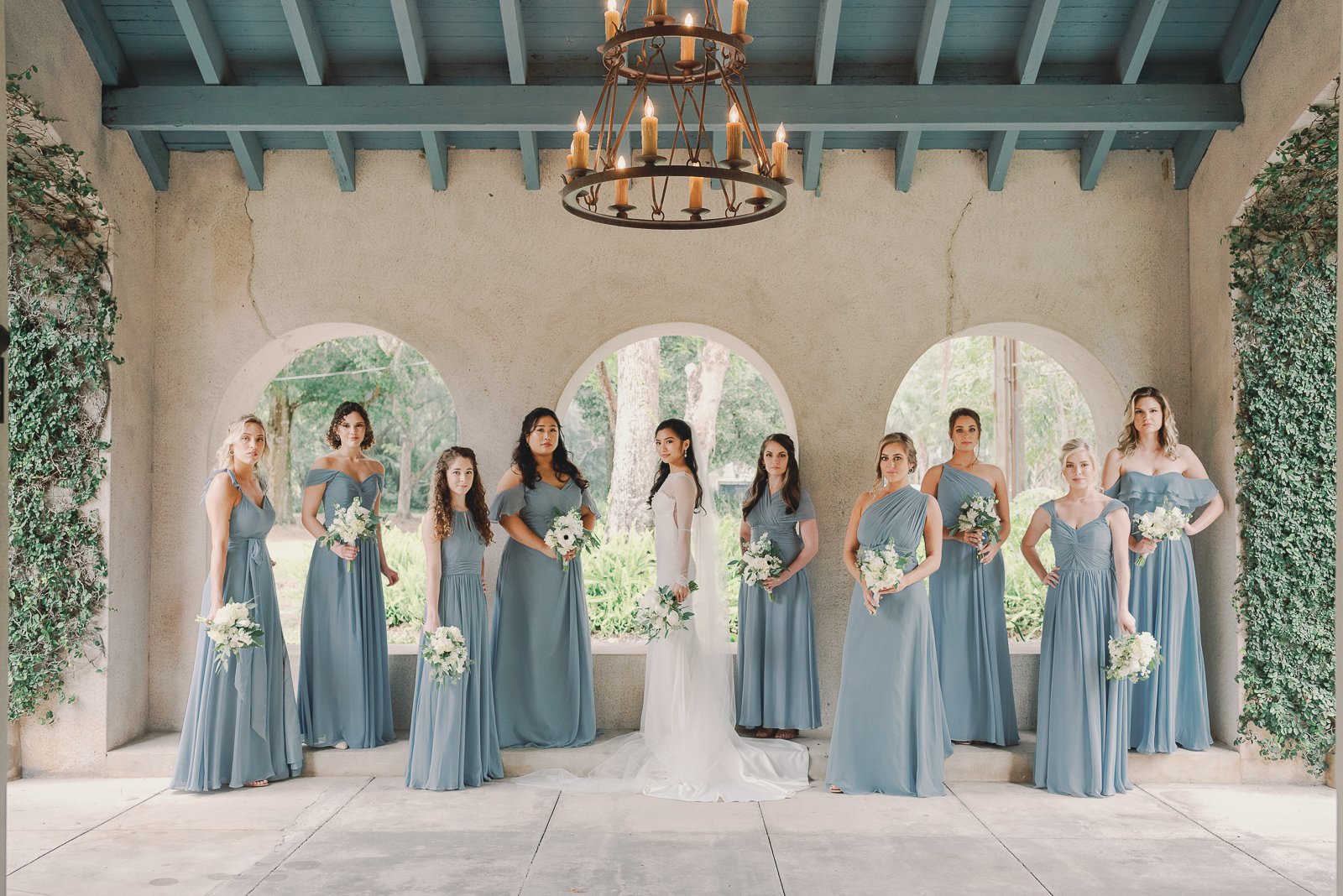 A beautiful bride surrounded by her bridal party of 9. She has 4 to her right and 5 to her left. All the women are holding delicate bouquets of flowers and the bridesmaids are dressed in long pale blue dresses.